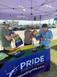 Volunteers hand out rainbow Pride Corpus Christi tote bags at a Pride Corpus Christi booth under a purple tent.