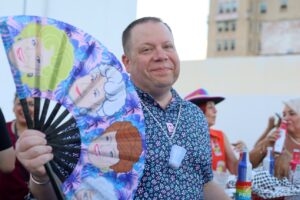 Pride Corpus Christi board member Eric Walls holds a Golden Girls fan at a downtown Pride pub crawl, with festive guests behind him.