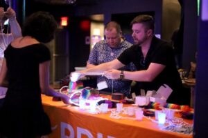 Pride board President Jonathan Swindle and Eric Walls hand out beads and rainbow Pride merchandise to a guest at a merch table during a downtown pub crawl.