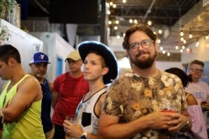 Attendees holding drinks at an indoor Pride pub crawl under string lights, one wearing a light-up cowboy hat.
