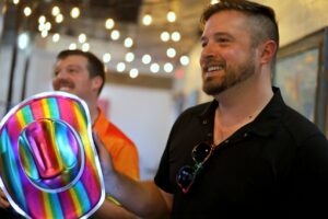 Pride board President Jonathan Swindle holds a rainbow cowboy hat at a Pride pub crawl, with a friend laughing under string lights.