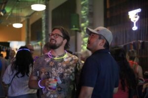 Two men with glow necklaces chat at a downtown Pride pub crawl, a neon cocktail sign glowing nearby.