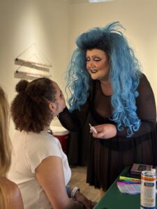 Ophelia Tits leans in to gently apply drag makeup to a smiling attendee at the Art of Drag K Space Contemporary event.