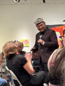 Emperor Raymond of TRE in a jeweled crown holds a microphone, greeting an applauding seated audience at a Pride event hosted by K Space Contemporary.