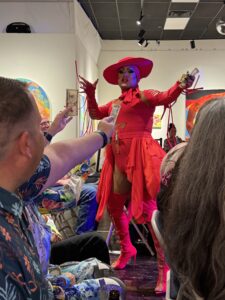 Drag performer in a red gown, hat, and pink boots collects tip dollars from an audience at an art gallery show.