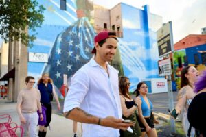 Smiling man in a backwards red cap leads a group on a downtown bar crawl past Pop's Bar mural.