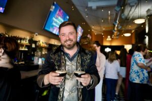 Jonathan Swindle in an embroidered jacket holds out two espresso martinis at a lively bar crawl event.