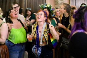 Costumed revelers laugh and dance at a medieval fantasy bar crawl, with flower crowns and a rainbow Pride sash.