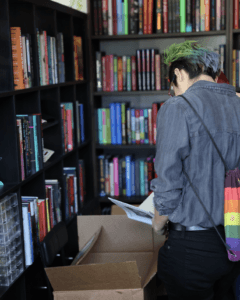 Person with green and blue hair and a rainbow Pride bag browses colorful books at a shelf.