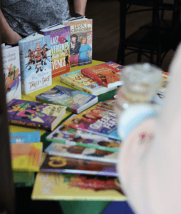 Display table of LGBTQ+ books including "Lucy, Censored" and "Bea Mullins Takes a Shot" at a Pride social.