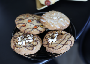 Four gourmet cookies topped with marshmallows, candy, and chocolate drizzle on a black plate at The Cookie Plot.