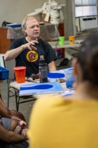 Instructor in a Buc-ee's shirt leads an art workshop, gesturing over blue-painted canvases and a heat gun.