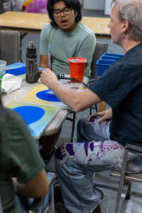 Teens gather around a table making blue-circle paintings at an Art Lab workshop, with a heat gun and paint supplies.