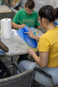 Teens painting at a paint-splattered table during an art lab, one holding a blue and yellow canvas.