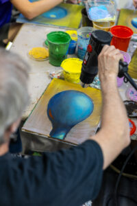 Artist uses a heat gun on a blue painting beside cups of colorful paint at an art workshop.