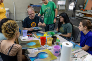 Instructor demonstrates painting to a group of teens around a table with paint cups and colorful canvases at an art lab.