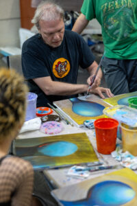 Person painting a blue orb on canvas at a community art table surrounded by paint cups and palettes.