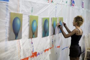 Teen artist in fishnet sleeves hangs a row of blue abstract paintings on a paint-splattered studio wall.