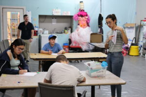 Instructor leads an art workshop as teens draw at tables, near a bin labeled "Clay & Tools.