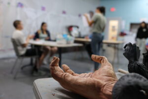 Sculpted hand and bird artworks in foreground as teens work and talk during an art lab session.