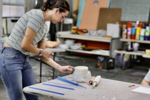 Teen uses a wire tool to cut a slab of clay in a colorful pottery studio during an art workshop.