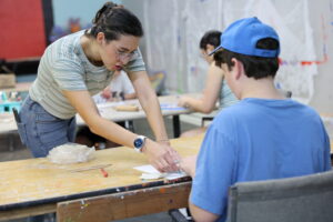 An instructor guides a teen through a clay project at a paint-splattered table during a hands-on art workshop.