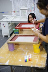 Two people smiling and decorating screen-printing frames with markers at a paint-splattered art studio table.