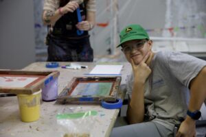 Smiling teen in a green John Deere cap at a paint-splattered table with screen-printing frames at an art lab.