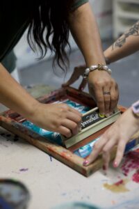 Hands pull a Speedball squeegee across a paint-splattered screen-printing frame at a teen art lab.