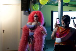 Brigitte Bandit in feather boa poses as attendees film on their phones at a Pride event.