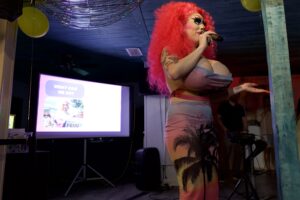 Brigitte Bandit speaks into a mic beside a "What Can We Do?" Pride presentation screen.