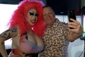 Brigitte Bandit with bright pink hair smiles for a selfie with a man in a floral shirt.