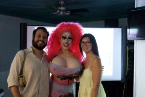 Brigitte Bandit with a bright pink wig poses with two smiling guests at an indoor event.