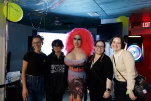 Drag performer Brigitte Bandit in a pink wig and sunset dress poses with four smiling guests at an indoor event.