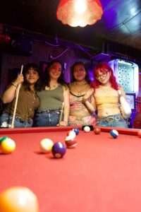 Four friends with pool cues smile behind a red billiards table at The Hidden Door with a Bullshooter dartboard.