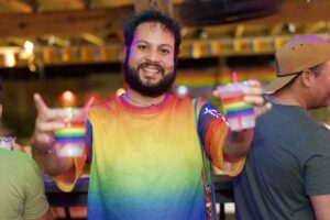 Orlando in a rainbow mesh jersey holds out two colorful drinks at the Pride kickoff party at The Hidden Door.