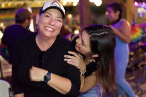 Two women embrace and smile warmly, one in a rainbow cap and the other in rainbow earrings, at the 2026 Pride kickoff event at The Hidden Door.