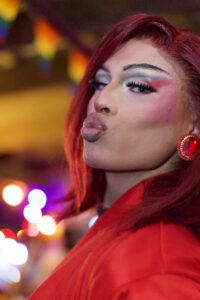 Drag performer with red hair and bold makeup blows a kiss beneath rainbow flags at Pride Kickoff.