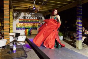 Drag performer twirls a flowing red gown onstage at The Hidden Door beneath a "Happy Pride" banner.