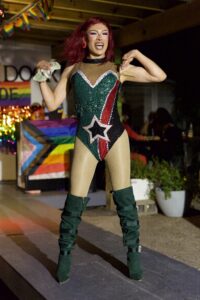 Drag performer in sequined star bodysuit holds tip money onstage by a Progress Pride flag at Pride Kickoff.