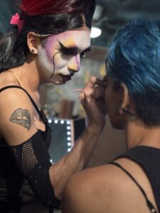 Drag performer in bold face makeup and a lip tattoo applies makeup to another artist at the Pride Corpus Christi makeup event.